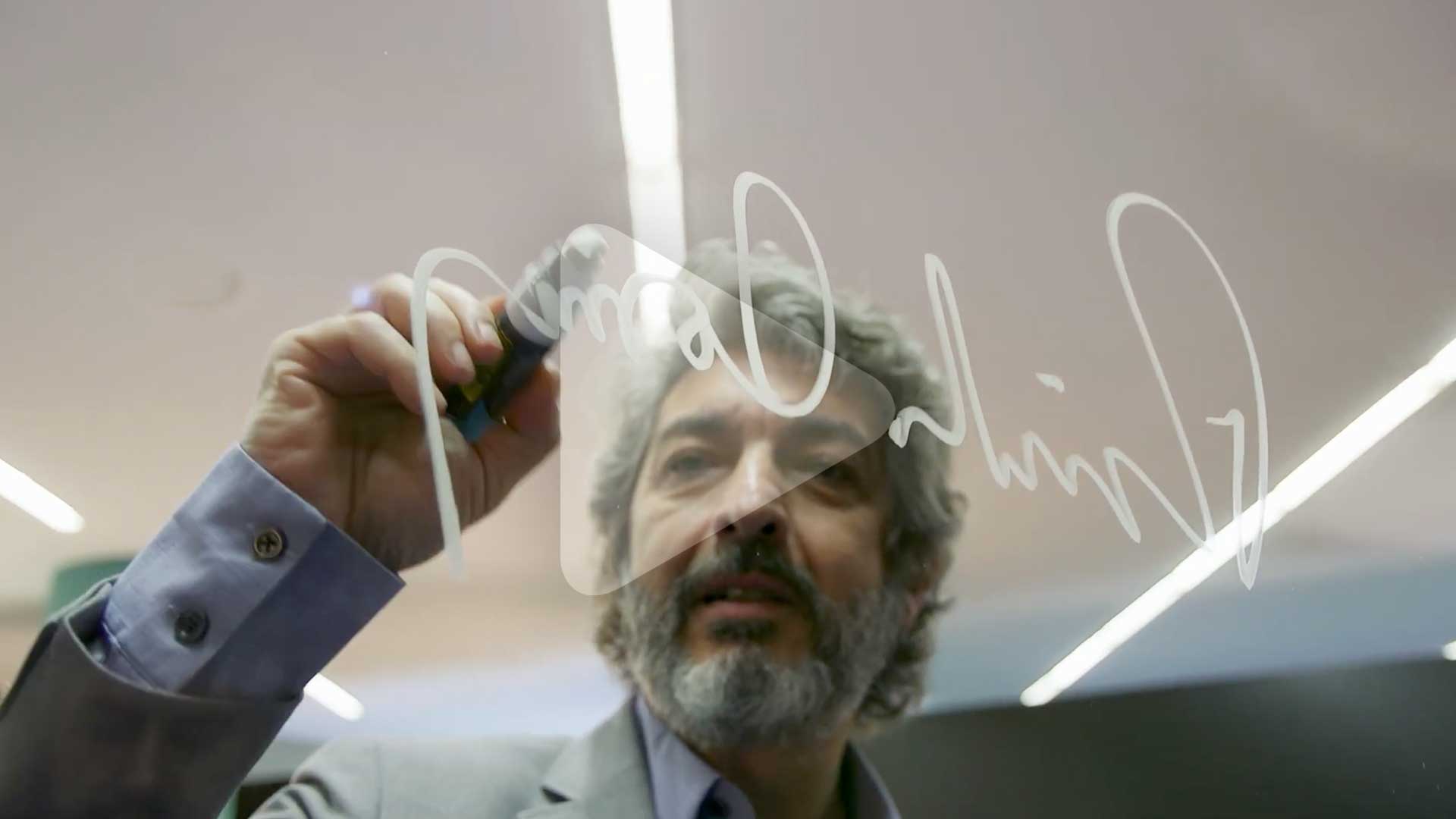 A man with gray hair and a beard is writing on a glass surface with a marker. He wears a suit and looks focused.