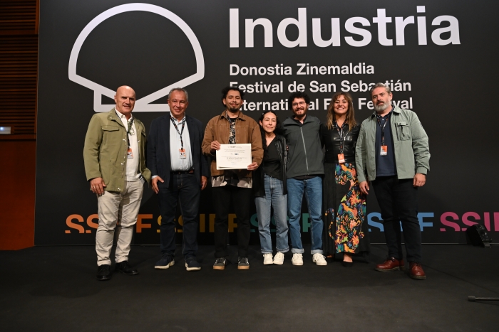 A group of six people stands on stage, holding an award certificate, with a backdrop featuring the text