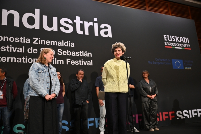 A group of people stands on stage at a film festival event, with one woman speaking into a microphone. The backdrop features festival branding.