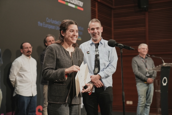 A woman smiles while holding a certificate on stage, accompanied by several men. A microphone is in front of her.